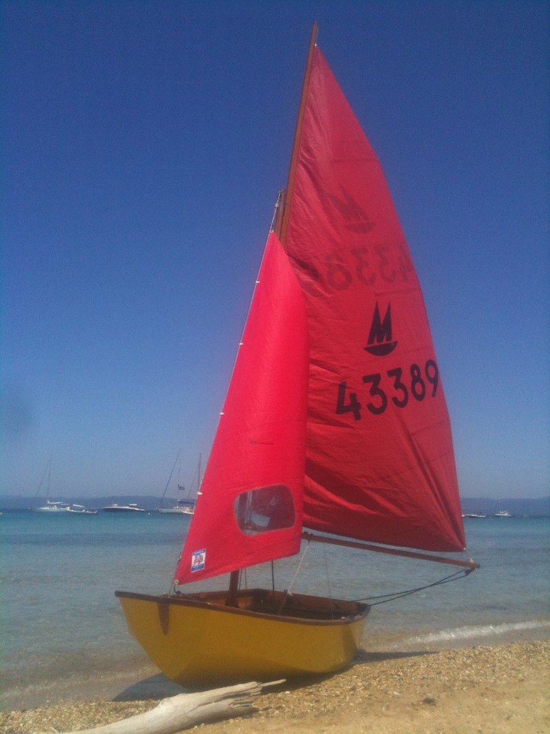 A yellow Mirror dinghy on a beach with cloudless blue sky and transparent blue water